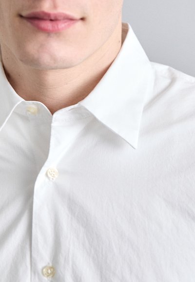 Camisa de algodón blanca de botones con cuello puntiagudo, que presenta una textura sutil y botones visibles a lo largo de la tapeta.