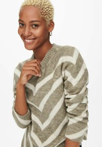 Pull en tricot beige et crème avec un motif en chevron ; il présente un col rond et des manches ajustées. Matière douce et texturée.
