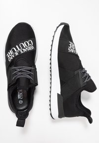 Svarta sportskor med ett slätt textilöverdrag, vit sula, elastiska snören och "Versace Jeans Couture" logotyp över vristen.