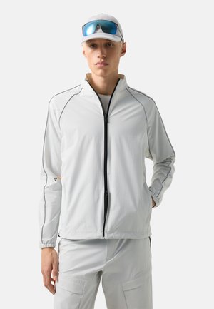 Jeune homme portant une veste zippée gris clair et un pantalon cargo, une casquette blanche avec des lunettes de soleil réfléchissantes bleues posées dessus, debout devant un fond uni.