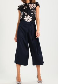 Czarna bluzka w kwiaty z jasnoróżowymi płatkami, z krótkim rękawem, w połączeniu z granatowymi szerokimi spodniami z wiązanym paskiem. Szare wysokie obcasy dopełniają stylizację.