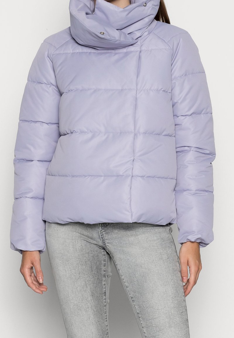 Femme portant une doudoune légère violette avec un col montant et un jean gris clair, debout devant un fond uni.