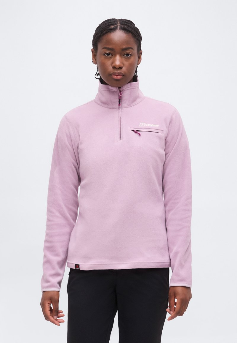 Γυναίκα που φοράει ένα ανοιχτό μωβ φλις με φερμουάρ μέχρι τη μέση και τσέπη στο στήθος, καθώς και μαύρο παντελόνι, στέκεται μπροστά σε λευκό φόντο.