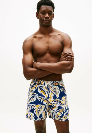 Muskulöser Mann mit verschränkten Armen, der marineblaue Badeshorts mit gelben und weißen tropischen Blattmustern trägt und vor einem einfarbigen Hintergrund steht.