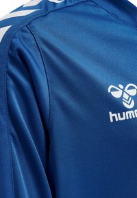 Maglia sportiva blu realizzata in tessuto morbido con logo bianco e accenti a zig-zag bianchi in contrasto lungo la spalla.