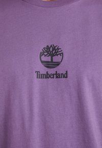 Violetinis medvilninis marškinėliai su juodu Timberland logotipu, turinčiu medžio grafiką, apvaliu dizainu ir įrengtu apykakle.