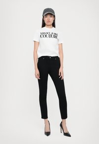 Balta medvilninė marškinėlių su užrašu "VERSACE JEANS COUTURE" juodu spalva, derinama su juodomis lieknomis kelnėmis ir juodais aukštakulniais bateliais. Pilkas kepuraitės akcentas.