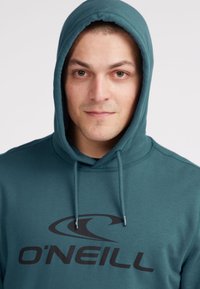 Sudadera color verde azulado hecha de una mezcla de algodón, con un bolsillo frontal, capucha ajustable y un gran logo negro "O'NEILL" en el pecho. Textura suave.