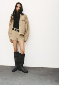 Beige corduroy-takki mustalla kauluksella, yhdistettynä beigeihin shortseihin ja mustiin polvisukkiin harmaalla tekstuuripinnalla valkoista taustaa vasten.