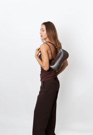 Bruine, gestructureerde, gebogen schoudertas gemaakt van een glanzend materiaal. Draagt op de rug, gecombineerd met een nauwsluitend topje en losse broek.