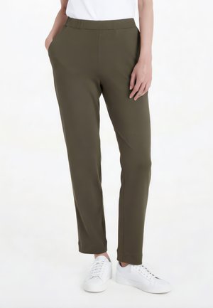 Persona che indossa pantaloni slim verde oliva con le mani in tasca e sneakers bianche, in piedi contro uno sfondo bianco.