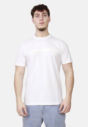 Uomo con capelli corti e ricci, che indossa una maglietta bianca a maniche corte e jeans celesti, in piedi contro uno sfondo chiaro.