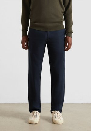 Navyblauwe broeken met een rechte snit en subtiele textuur, gecombineerd met crèmekleurige sneakers. De outfit wordt aangevuld met een olijfgroen trui.