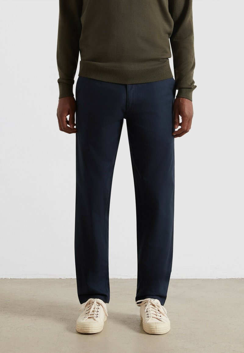 Navyblauwe broeken met een rechte snit en subtiele textuur, gecombineerd met crèmekleurige sneakers. De outfit wordt aangevuld met een olijfgroen trui.