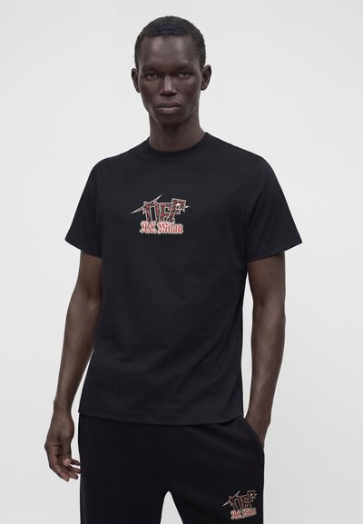 Homme portant un T-shirt noir et un pantalon avec les logos rouges et blancs Off-White AC Milan, debout devant un fond gris uni.