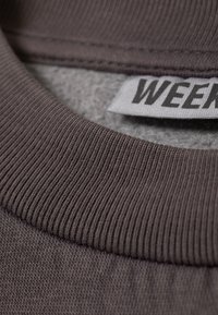 Dunkelbrauner gerippter Stoff mit einem strukturierten Kragen und einem Etikett, auf dem "WEEK" in fetter schwarzer Schrift steht.