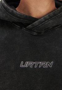Sudadera negra de tela suave, con capucha ajustable y gran logo blanco "URTRN" en el frente. Textura desgastada visible.