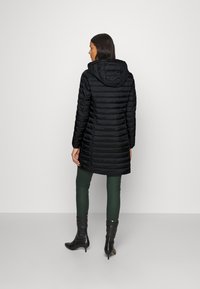 Manteau d'hiver noir avec capuche, présentant un design matelassé et des manches longues. Porté avec un legging vert et des bottines noires à hauteur de cheville.