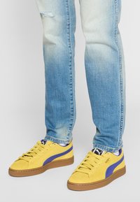 Żółte zamszowe sneakersy z niebieskim paskiem, z teksturowaną gumową podeszwą i klasycznym sznurowaniem, w połączeniu z jasnoniebieskimi dżinsami.