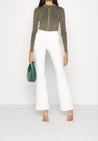 Top olivo de manga larga con costillas, pantalones acampanados blancos de tiro alto y un bolso verde con textura. Sandalias claras con tiras completan el conjunto.