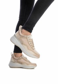 Zapatillas deportivas beige con parte superior texturizada, diseño de cordones, detalles en dorado y suela de goma blanca, combinadas con jeans negros.