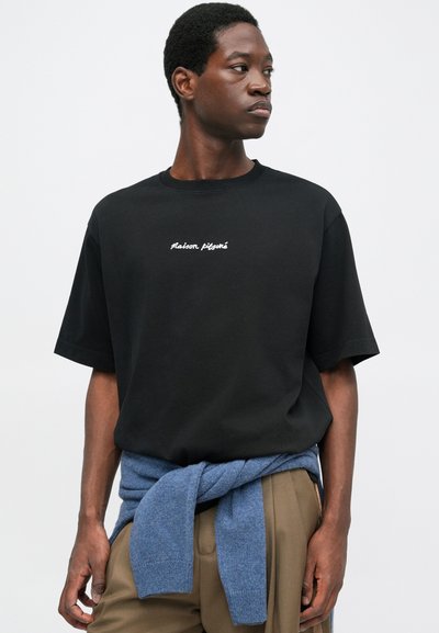 Schwarzes Baumwoll-T-Shirt mit kurzen Ärmeln, welches auf der Brust weißen, gestickten Text "Maison Kitsuné" trägt. Ein blauer Strickpullover ist um die Taille gebunden.