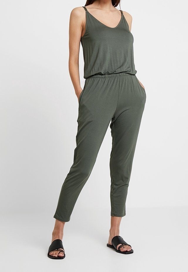 Woman wearing olive green sleeveless jumpsuit with elastic waist and black slide sandals standing against white background.