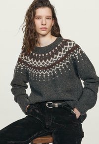 Grauer Wollpullover mit einem gemusterten Einsatz in Creme und Weinrot, Rundhalsausschnitt und langen Ärmeln, kombiniert mit schwarzen Cordhosen.