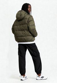 Veste matelassée verte avec capuche et texture en relief. Portée avec un pantalon cargo noir et des chaussures de sport blanches avec des accents noirs.