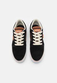 Black suede sneakers with cream laces and orange accents, featuring a padded tongue and textured side panel. Rubber sole with white trim.