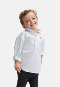 Camisa de botones a rayas azul claro con mangas remangadas, combinada con pantalones oscuros; presenta un pequeño logo bordado en el pecho.