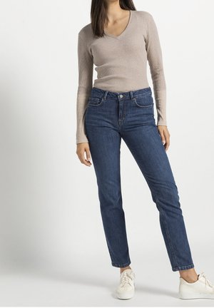 Femme portant un haut beige à manches longues avec un col en V, un jean bleu taille haute et des baskets blanches, debout devant un fond uni.