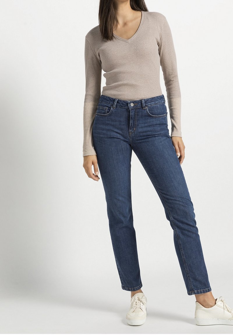 Femme portant un haut beige à manches longues avec un col en V, un jean bleu taille haute et des baskets blanches, debout devant un fond uni.