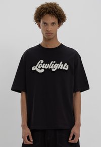 Jauns vīrietis ar cirtainiem matiem, ģērbies melnā oversize T-kreklā ar baltiem teksturētiem burtiem, kuros rakstīts "Lowlights".