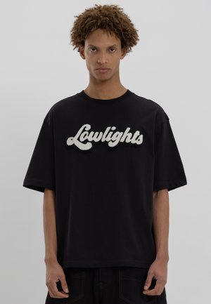 Fiatalember, göndör hajjal, fekete, oversized pólót visel, melyen fehér, texturált betűtípussal "Lowlights" felirat található.