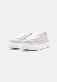 Μπεζ σουέντ αθλητικά παπούτσια με λευκές λαστιχένιες σόλες, διαθέτοντας λευκά κορδόνια και ραμμένα διακοσμητικά στη πάνω πλευρά. Μινιμαλιστικός σχεδιασμός, στρογγυλεμένο σχήμα.