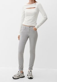 Top blanco de manga larga con un corte central, combinado con jeans gris claro ajustados y zapatillas grises con acentos negros.
