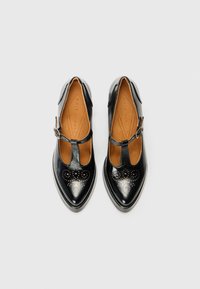 Chaussures T-strap en cuir noir avec bouts pointus, perforations décoratives sur le bout, et une fermeture à boucle à la cheville.