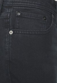 Los jeans de mezclilla oscura presentan un bolsillo delantero con un botón negro, un remache plateado y una tela texturada, mostrando un diseño suave y clásico.