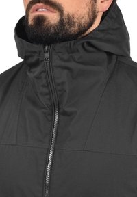 Veste noire à capuche avec une fermeture éclair intégrale, présentant une texture lisse et des coutures contrastantes. Le tissu semble résistant à l'eau et durable.