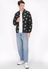 Zwarte bomberjack met kleurrijke palmenpatronen, gecombineerd met een eenvoudig wit T-shirt en lichtblauwe spijkerbroek, gedragen met witte sneakers.