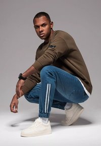 Olivgrön sweatshirt med ribbade muddar, blå denimjoggers med vita ränder och vita sneakers. Figuren hukar sig och visar upp vardagskläder.