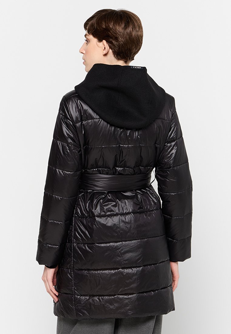 Kobieta ubrana w błyszczący czarny puchowy płaszcz z paskiem i dużym czarnym kapturem, widziana z tyłu na tle jasnego, jednolitego tła.