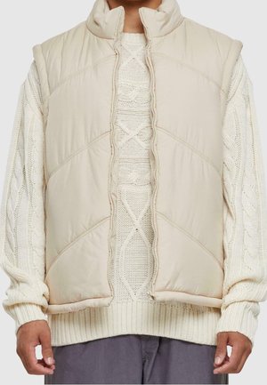 Personne portant un pull crème en maille torsadée sous une veste sans manches matelassée beige avec un col montant, associé à un pantalon gris.