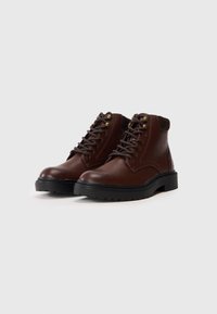 Bottes en cuir marron jusqu'à la cheville avec des bouts ronds, semelles en caoutchouc noir et œillets en métal pour les lacets. Présente un panneau arrière surélevé marron.