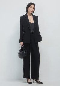 Donna che indossa un completo nero con righe bianche, blusa nera, tiene una borsa nera ed è in tacchi a punta neri, in piedi su uno sfondo bianco.