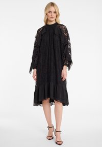 Schwarzes Spitzenkleid mit Rüschensäumen und gestuftem Saum. Verfügt über einen hohen Halsausschnitt und transparente Akzente. Kombiniert mit schwarzen Riemchensandalen.