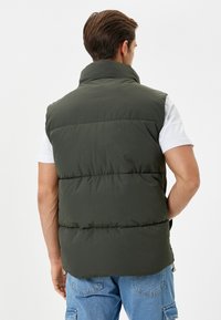 Chaleco acolchado verde oliva con alto cuello, costuras horizontales y un ajuste relajado, llevado sobre una camiseta blanca y pantalones cortos de mezclilla.