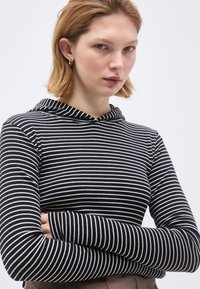 Noor naine lühikeste blondide juustega, kannab kuldseid rõngaskõrvarõngaid ja musta-valge triibulist kapuutsiga pika varrukaga topi, käed rüpes.