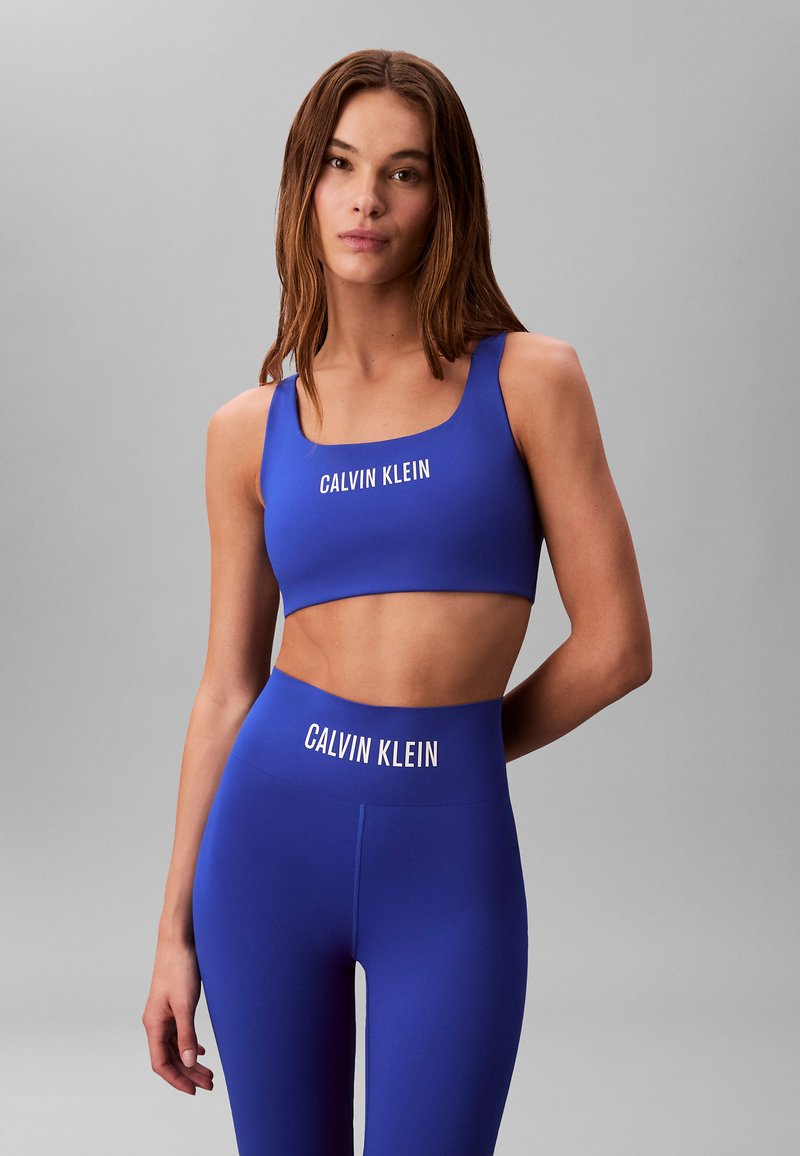 Niebieski sportowy crop top i legginsy z wysokim stanem, z białym logo "CALVIN KLEIN", wykonane z elastycznego materiału, o gładkiej fakturze.